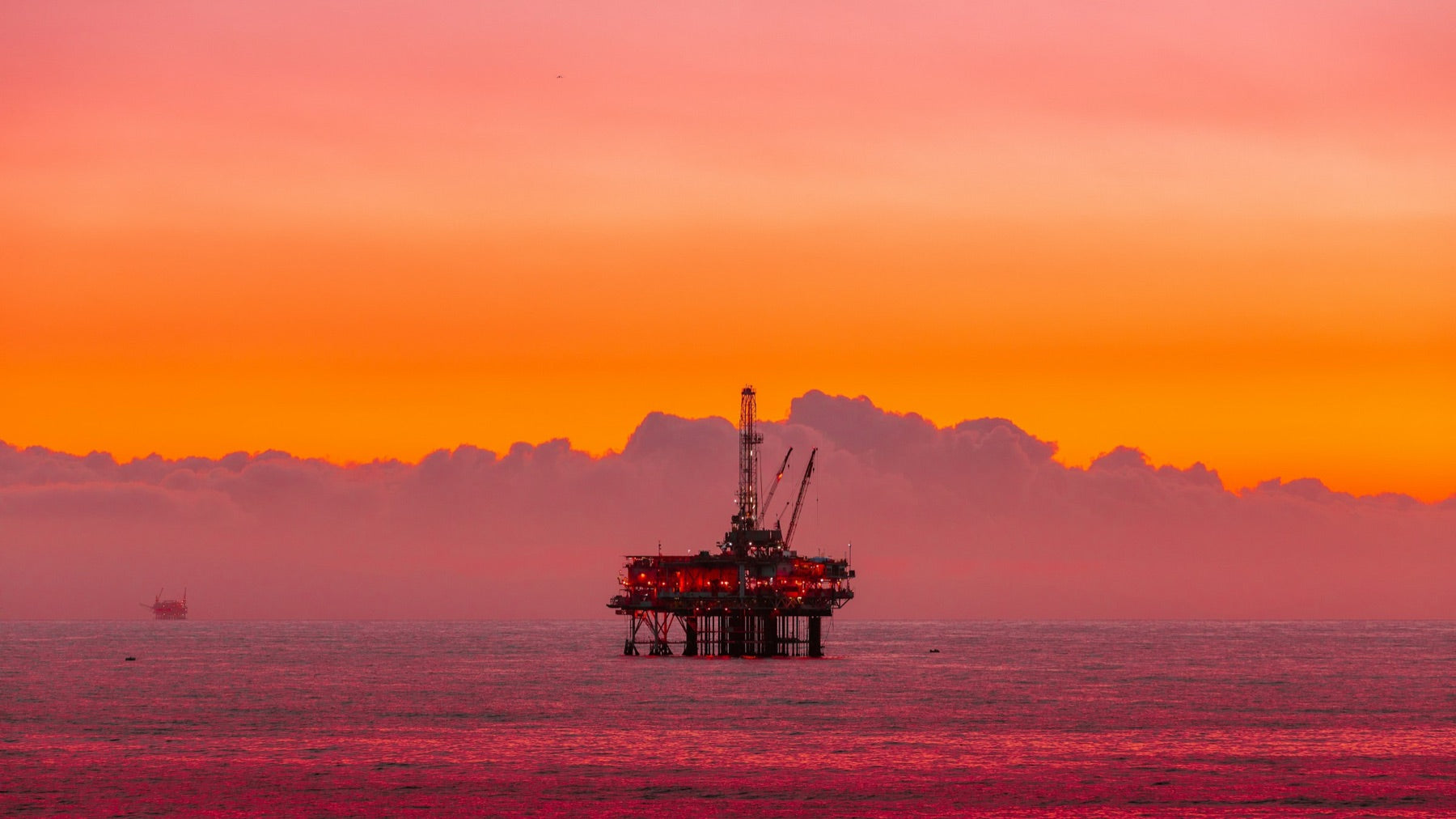 An offshore oil platform silhouetted against a vivid orange and pink sunset over the ocean